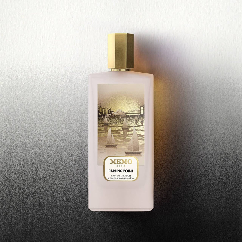 A pale pink rectangular perfume bottle with a gold cap and label featuring an illustration of sailboats on water, labeled ’memo paris darling point.’