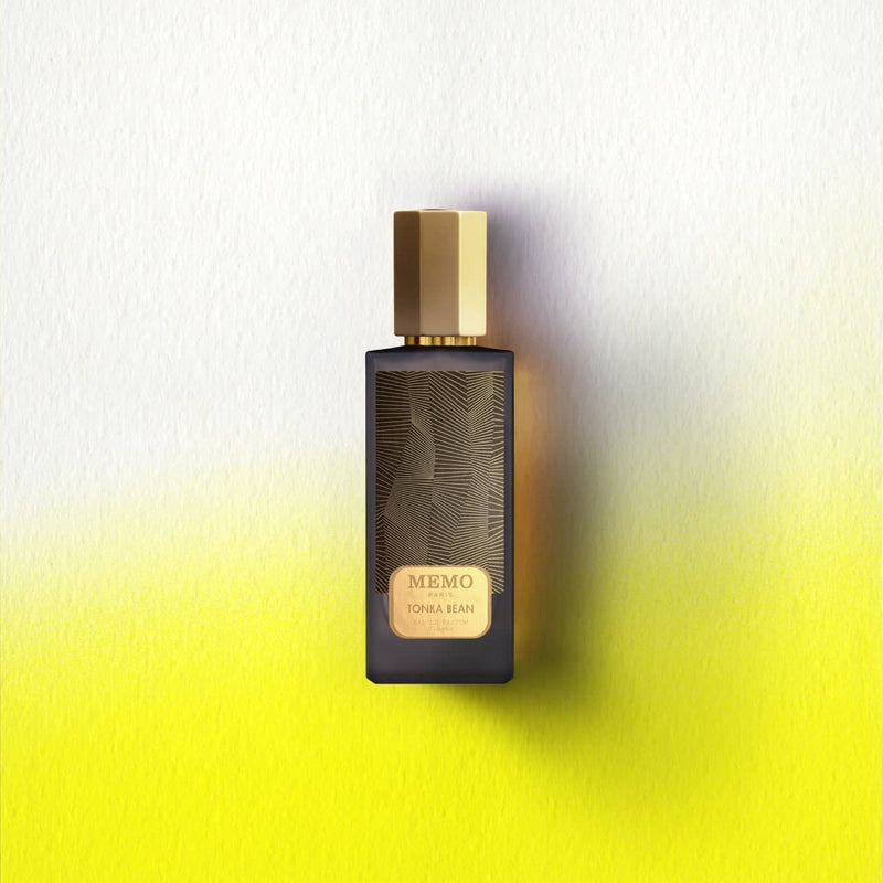 A sleek black glass perfume bottle with a gold hexagonal cap and intricate golden leaf patterns, labeled ’memo paris tonka bean.’